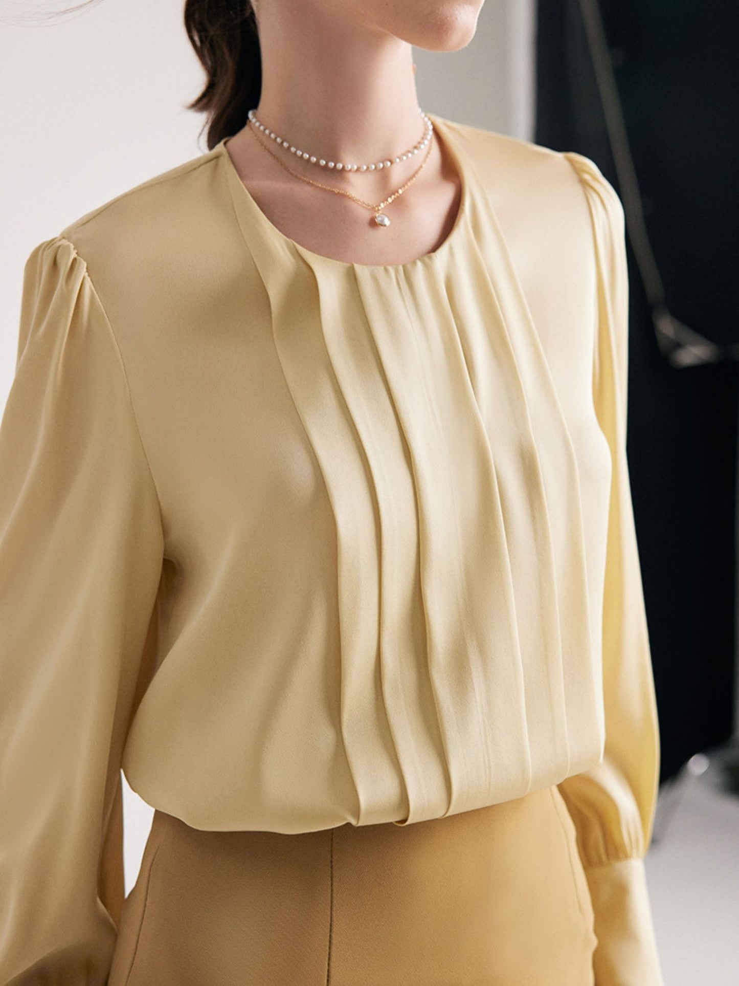 A woman wearing a round neck silk blouse with pleated details at the front, long sleeves, and buttoned cuffs, in khaki.