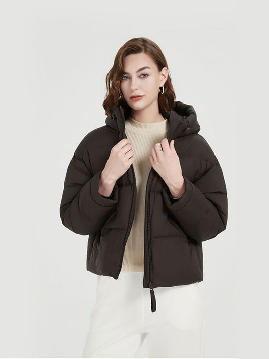 A female model wearing a black thick down jacket with a hood and functional pockets holding the collar of the jacket.