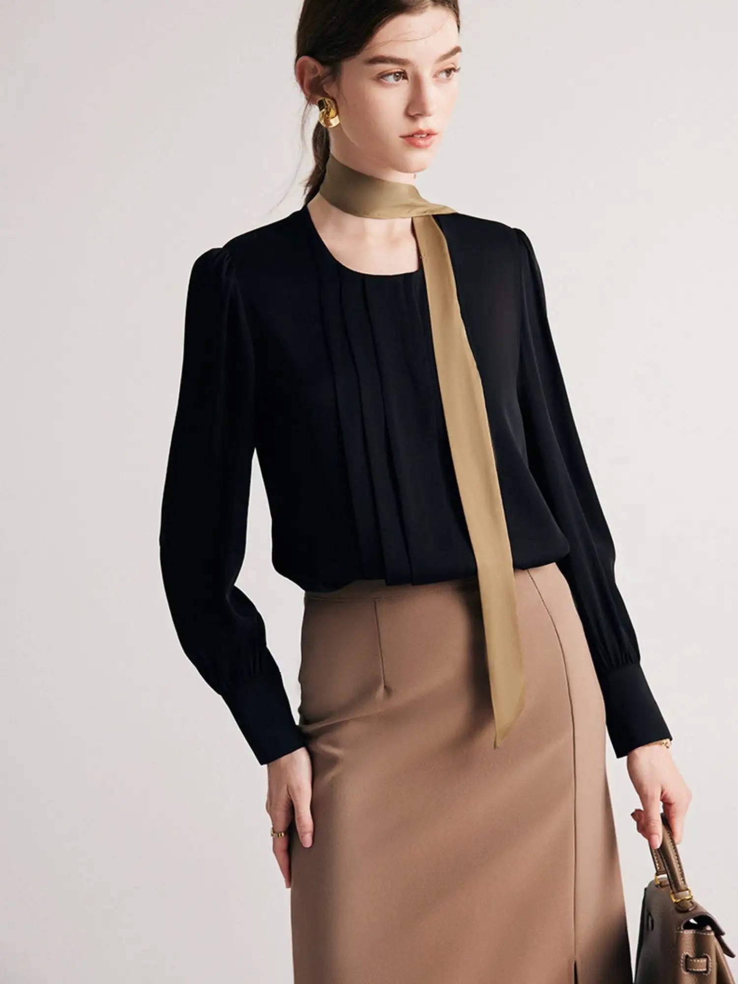 A woman wearing a round neck silk blouse with pleated details at the front, long sleeves, and buttoned cuffs, in black.