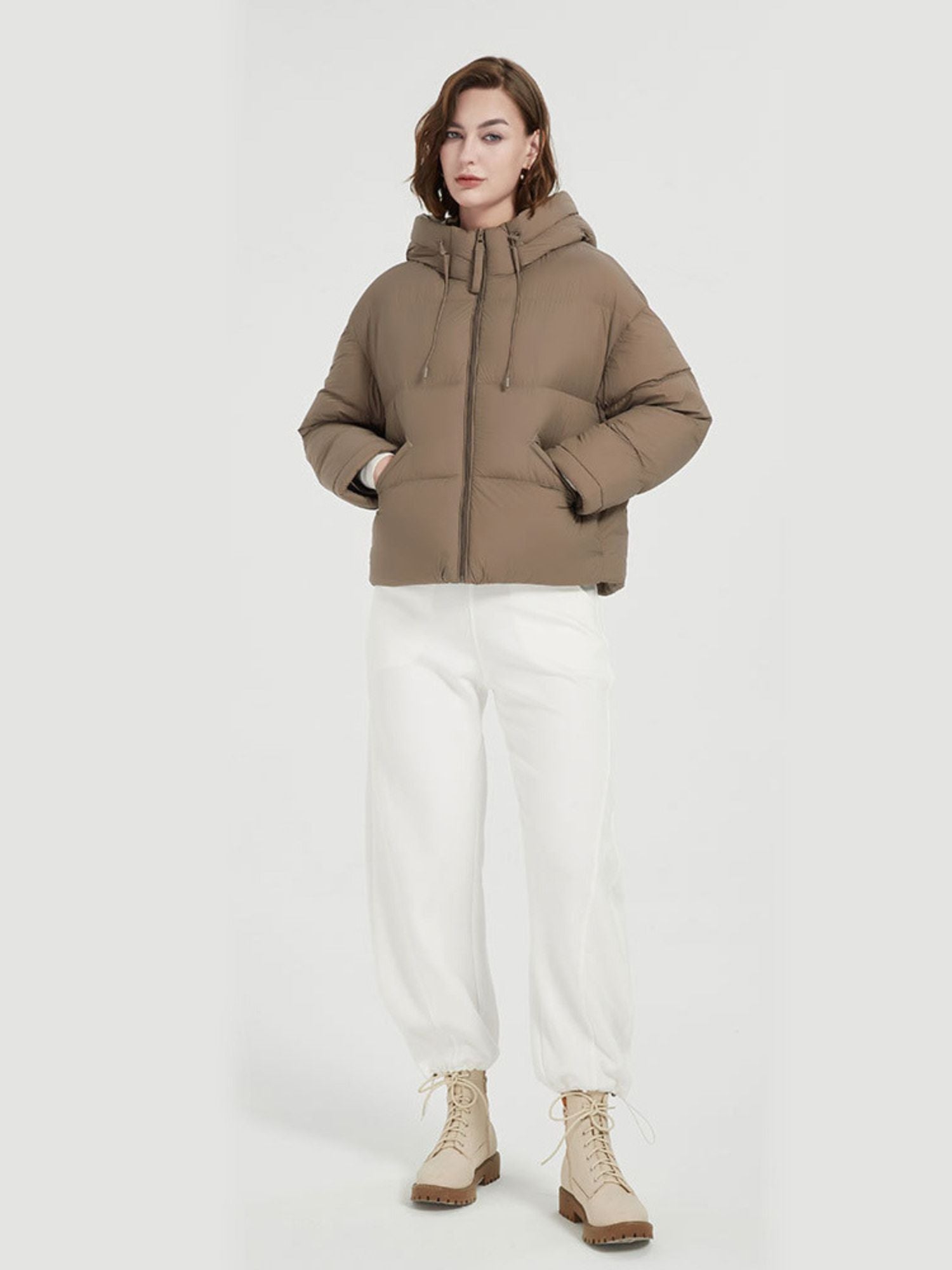 A female model wearing a brown thick down jacket with a hood and functional pockets, paired with white pants.